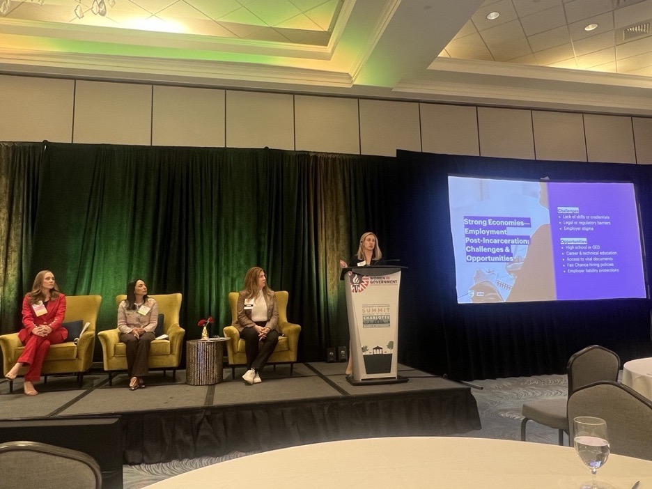 Megan speaking in a podium, and 3 other women in the pannel